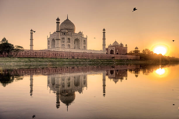 The early morning light bathes the Taj Mahal in a warm glow, highlighting its ivory-white marble and reflecting beautifully in the calm waters of the Yamuna River.