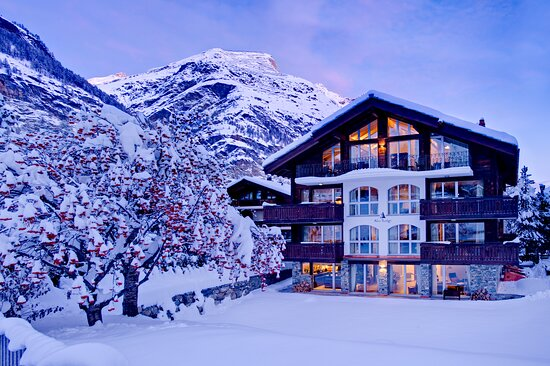 Cozy Swiss mountain lodge surrounded by snowy peaks and pine trees, with smoke rising from a chimney and warm lighting inside